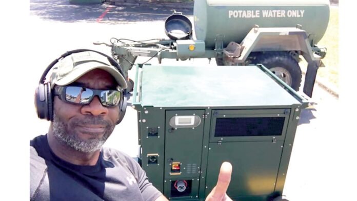 Moses West, usando fones de ouvido e óculos de sol, dando um joinha ao lado de seu Atmospheric Water Generator (AWG), com um tanque de água potável ao fundo.